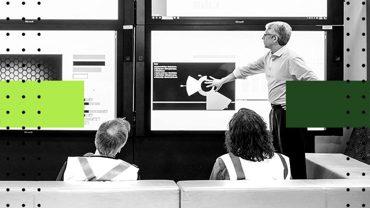 Two individuals in safety vests listen to a presentation while a person points at a large screen displaying various content.