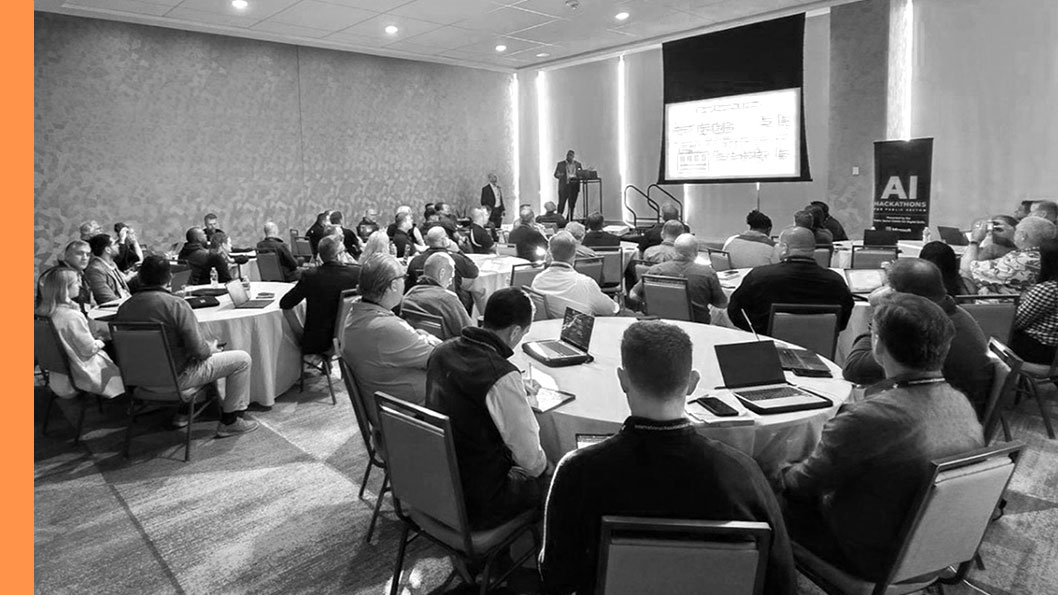 A conference room filled with people sitting around tables, listening to a speaker presenting information at the front of the room