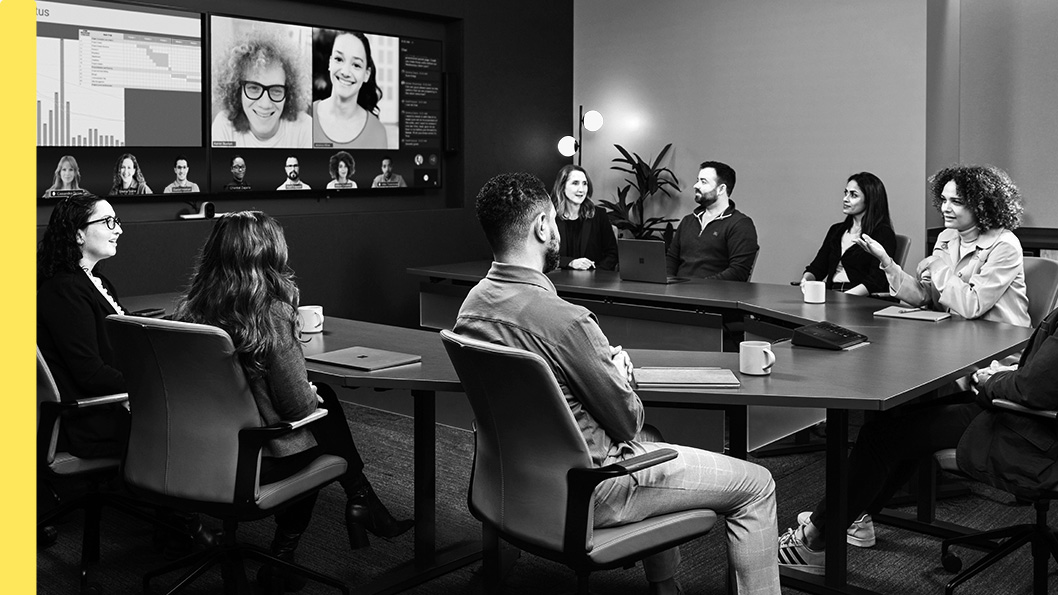 A conference room with people seated at a table, participating in a video conference displayed on large screens.
