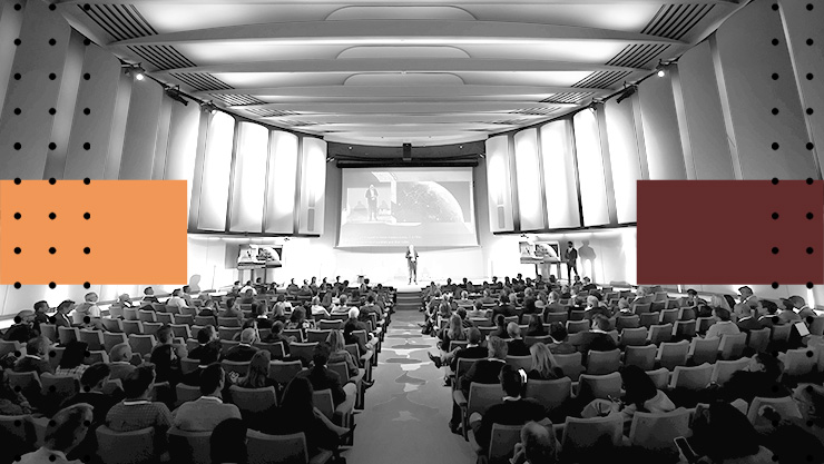 A wide view of a large auditorium filled with seated attendees facing a stage and presentation screen.