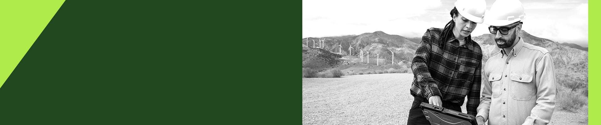 Two people wearing hard hats review information on a rugged laptop at an outdoor work site with wind turbines in the background.