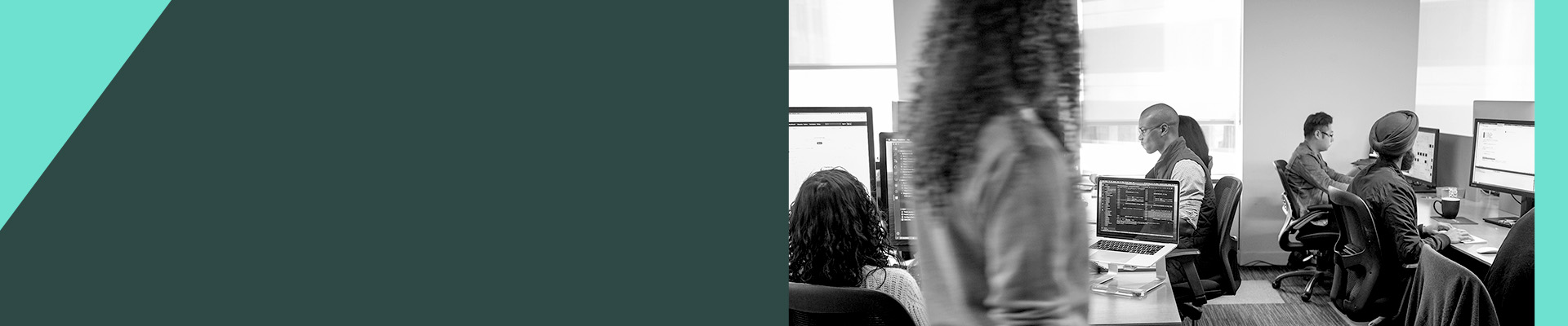 People working at computers in a shared office workspace, with multiple desks and monitors visible.