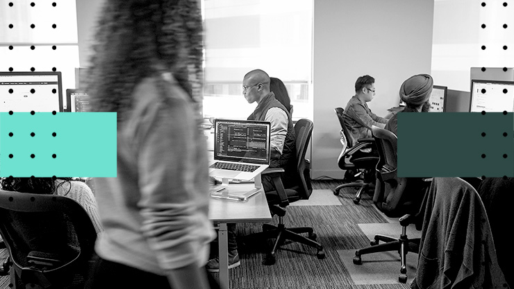 People working at computers in a shared office workspace, with multiple desks and monitors visible.
