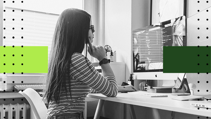 Person with long hair sits at a desk using a desktop computer, hand resting against the face in a workspace.